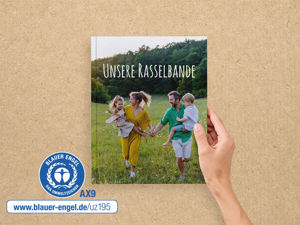 Eine Hand hält ein A4 Hoch Fotobuch. Auf dem Cover ist eine Familie auf einer Wiese abgebildet. Daneben ist der Blaue Engel.