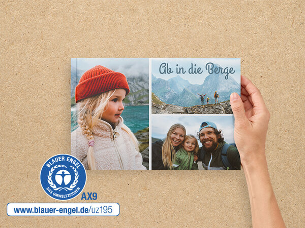 Eine Hand hält ein A4 Quer Fotobuch im. Auf dem Cover ist ein Familie beim Wandern abgebildet. Daneben ist der Blaue Engel.